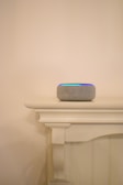 A smart speaker with a fabric covering sits atop a painted wooden mantel. The mantel features clean, simple lines and a decorative corbel underneath. Soft lighting casts a gentle shadow on the neutral-colored wall in the background.
