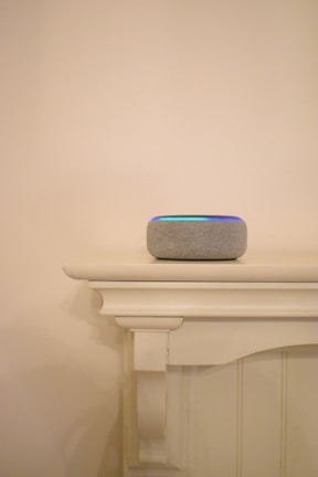 A smart speaker with a fabric covering sits atop a painted wooden mantel. The mantel features clean, simple lines and a decorative corbel underneath. Soft lighting casts a gentle shadow on the neutral-colored wall in the background.