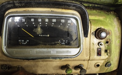 An aged and weathered car dashboard with visible speedometer and fuel gauges. The surface is covered in dirt and rust, and the panel displays a mix of analog dials and buttons. The dashboard's color is predominantly a faded green with patches of rust and grime.