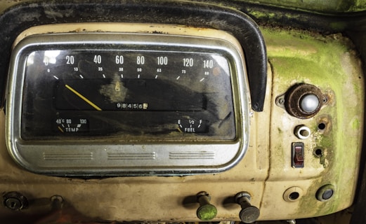 An aged and weathered car dashboard with visible speedometer and fuel gauges. The surface is covered in dirt and rust, and the panel displays a mix of analog dials and buttons. The dashboard's color is predominantly a faded green with patches of rust and grime.
