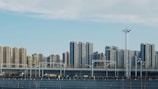 A cityscape featuring a series of high-rise apartment buildings under a partly cloudy sky. In the foreground, there is an elevated railway track with electrical poles and infrastructure. The scene reflects urban development and modern architecture.