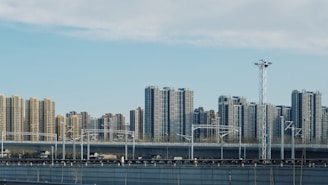 A cityscape featuring a series of high-rise apartment buildings under a partly cloudy sky. In the foreground, there is an elevated railway track with electrical poles and infrastructure. The scene reflects urban development and modern architecture.