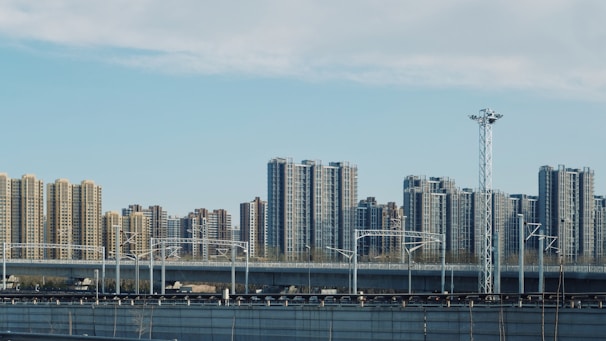 A cityscape featuring a series of high-rise apartment buildings under a partly cloudy sky. In the foreground, there is an elevated railway track with electrical poles and infrastructure. The scene reflects urban development and modern architecture.