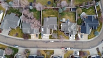 Aerial view of a suburban neighborhood featuring several well-arranged houses. Each house has its own driveway and lawn, with some cars parked outside. The properties are separated by trees and fences, and the streets are curvaceous and lined with sidewalks.