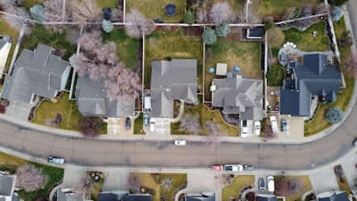 Aerial view of a suburban neighborhood featuring several well-arranged houses. Each house has its own driveway and lawn, with some cars parked outside. The properties are separated by trees and fences, and the streets are curvaceous and lined with sidewalks.