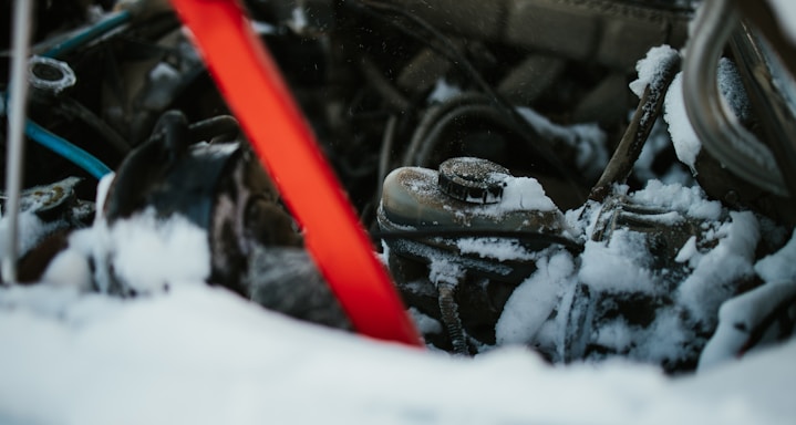 A close-up view of a snow-covered car engine with visible parts like a radiator cap and various wires. A red diagonal object, possibly a support bar, contrasts against the dark, snow-dusted machinery.