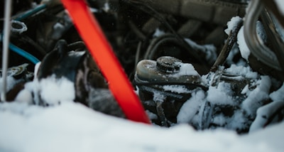 A close-up view of a snow-covered car engine with visible parts like a radiator cap and various wires. A red diagonal object, possibly a support bar, contrasts against the dark, snow-dusted machinery.
