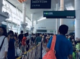 A bustling train station with a diverse crowd of people walking through the concourse. Large pillars and high ceilings are present, with directional signage in both Chinese and English. The atmosphere is busy and somewhat chaotic with people carrying luggage and wearing casual attire.