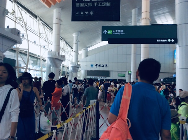 A bustling train station with a diverse crowd of people walking through the concourse. Large pillars and high ceilings are present, with directional signage in both Chinese and English. The atmosphere is busy and somewhat chaotic with people carrying luggage and wearing casual attire.