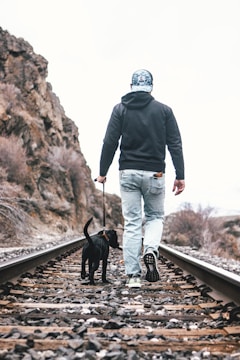 A person in casual clothing walks along a railway track holding a leash attached to a small black dog. The setting appears to be outdoors with rocky terrain and sparse vegetation visible on one side. The sky is cloudy, giving a muted atmosphere to the scene.