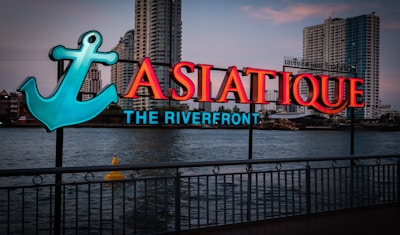 A neon sign with the text 'ASIATIQUE THE RIVERFRONT' is prominently displayed along a waterfront. The sign includes a large teal anchor graphic on the left. In the background, tall buildings are visible across the river at dusk.