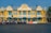 A bright yellow colonial-style building with blue shutters and decorative white accents stands prominently. In front of the building, a row of colorful tuk-tuks lined up along the street. Street lamps and barriers are also visible, along with some greenery to the right.