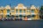 A bright yellow colonial-style building with blue shutters and decorative white accents stands prominently. In front of the building, a row of colorful tuk-tuks lined up along the street. Street lamps and barriers are also visible, along with some greenery to the right.