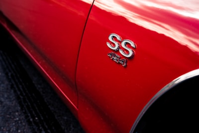 A close-up view of the side of a red car displaying chrome 'SS 454' badging, reflecting light and casting soft highlights.