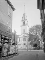 A black and white photograph of a street scene with a prominent church steeple in the background. The church building features classical architecture with a tall spire. In the foreground, there are several vintage movie posters on a wooden structure, with details of the films visible. The setting is framed by buildings on either side and trees adding a touch of greenery.