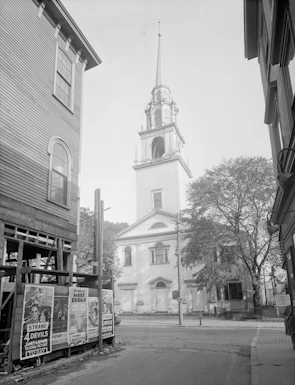 A black and white photograph of a street scene with a prominent church steeple in the background. The church building features classical architecture with a tall spire. In the foreground, there are several vintage movie posters on a wooden structure, with details of the films visible. The setting is framed by buildings on either side and trees adding a touch of greenery.