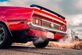 A classic red muscle car with a prominent rear spoiler is parked on a road. The car has chrome accents and distinctive rear lights, with a visible license plate at the back. A trail of exhaust fumes is visible, suggesting the car might be running. The background features an overcast sky and dry vegetation.