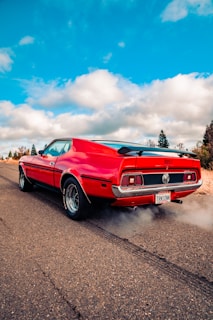 A classic red muscle car is parked on an open road, releasing smoke from the exhaust. The concrete road stretches into the distance under a bright blue sky with scattered white clouds. Surrounding the road are trees and dry grass, suggesting a rural setting.