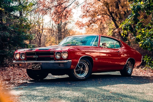 A classic red muscle car with the SS emblem is parked on a road surrounded by autumn foliage. The car's sleek body and chrome details are highlighted by the natural lighting, creating a nostalgic atmosphere.