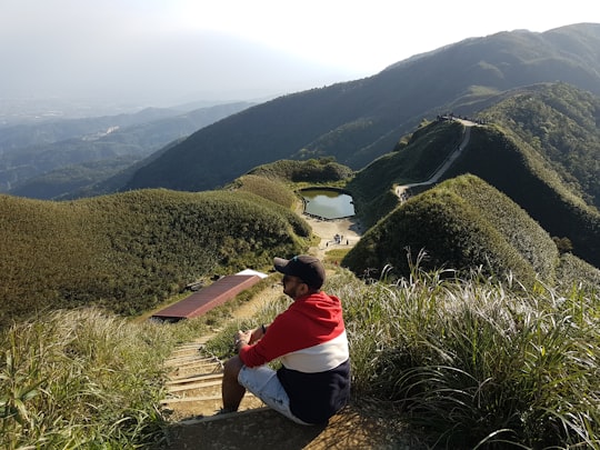 person in red and white hoodie sitting on brown rock looking at green mountains during daytime in Jiaoxi Township Taiwan