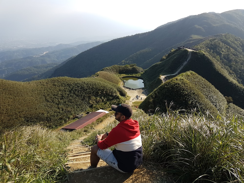 person in red and white hoodie sitting on brown rock looking at green mountains during daytime