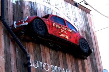 A red vintage car is mounted vertically on a wooden wall with the text 'Mechanic on Duty' displayed on the side. The car appears weathered, adding a rustic charm to the installation. The wall features additional text below, partially visible, displaying 'AUTO REPAIR'.