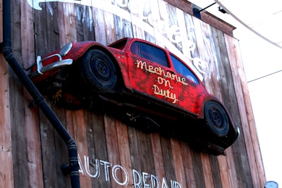A red vintage car is mounted vertically on a wooden wall with the text 'Mechanic on Duty' displayed on the side. The car appears weathered, adding a rustic charm to the installation. The wall features additional text below, partially visible, displaying 'AUTO REPAIR'.