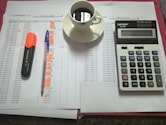 A workspace setting with financial documents, a black calculator displaying numbers, a cup of black coffee on a saucer, an orange highlighter, and a blue pen on a table.