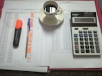 A workspace setting with financial documents, a black calculator displaying numbers, a cup of black coffee on a saucer, an orange highlighter, and a blue pen on a table.