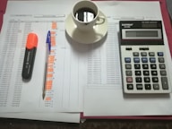 A workspace setting with financial documents, a black calculator displaying numbers, a cup of black coffee on a saucer, an orange highlighter, and a blue pen on a table.
