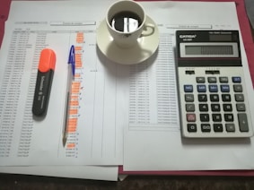 A workspace setting with financial documents, a black calculator displaying numbers, a cup of black coffee on a saucer, an orange highlighter, and a blue pen on a table.