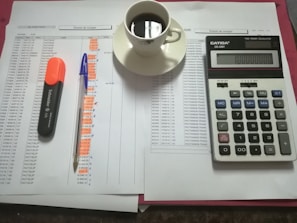 A workspace setting with financial documents, a black calculator displaying numbers, a cup of black coffee on a saucer, an orange highlighter, and a blue pen on a table.