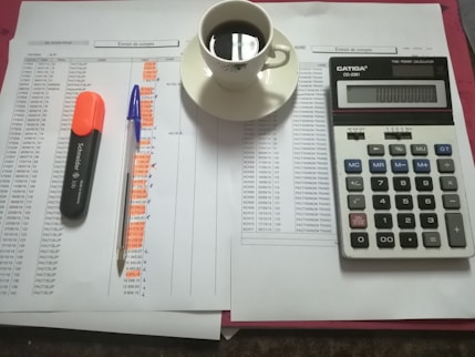 A workspace setting with financial documents, a black calculator displaying numbers, a cup of black coffee on a saucer, an orange highlighter, and a blue pen on a table.