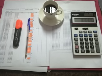 A workspace setting with financial documents, a black calculator displaying numbers, a cup of black coffee on a saucer, an orange highlighter, and a blue pen on a table.
