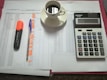 A workspace setting with financial documents, a black calculator displaying numbers, a cup of black coffee on a saucer, an orange highlighter, and a blue pen on a table.