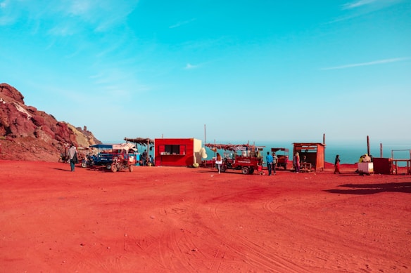 A red cliffside with several small, rustic wooden and metal structures sits above a vast blue sky and sea. People gather around these makeshift stalls near vehicles, giving a sense of a remote market or resting spot near a rocky landscape.