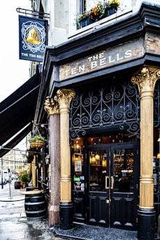 A historic pub with ornate architectural details features prominently. The entrance showcases intricate wrought ironwork and grand golden columns. The sign above the door reads 'The Ten Bells' in aged, rustic lettering. The facade includes floral decorations, and the street setting has a classic, old-world ambiance.