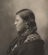 A sepia-toned portrait of a person in profile, showcasing long, dark hair and wearing large circular earrings. The individual is dressed in a patterned garment and wrapped in a dark fabric. The background is a soft, blurred gradient.