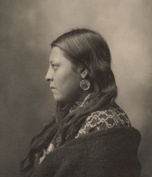 A sepia-toned portrait of a person in profile, showcasing long, dark hair and wearing large circular earrings. The individual is dressed in a patterned garment and wrapped in a dark fabric. The background is a soft, blurred gradient.