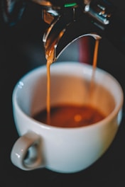 A ceramic cup with a handle is positioned under a coffee machine spout, capturing rich, dark espresso as it streams down. The focus is on the coffee flowing into the cup, creating a sense of warmth and comfort.