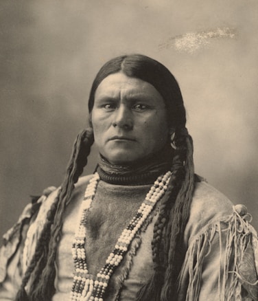A sepia-toned portrait of a person with long braided hair. They wear a high-necked garment and are adorned with several layers of beaded necklaces. Their expression is serious and contemplative, capturing a moment of stillness.
