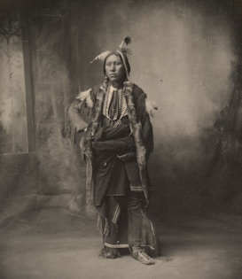 A black and white portrait of a person dressed in traditional attire. The clothing is adorned with feathers, fur, and intricate patterns, suggesting cultural significance. The background is blurred, focusing attention on the individual.