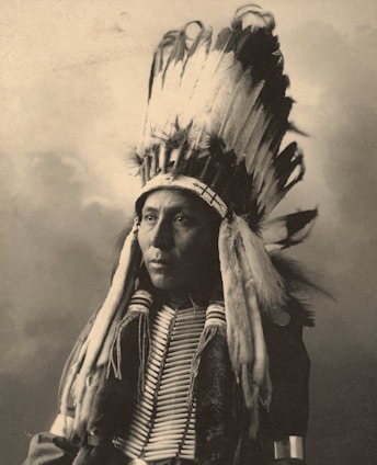 A person wearing a traditional Native American headdress with long feathers. The attire includes decorative elements and a beaded necklace. The image has a sepia tone, giving it an antique appearance.