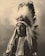 A person wearing a traditional Native American headdress with long feathers. The attire includes decorative elements and a beaded necklace. The image has a sepia tone, giving it an antique appearance.