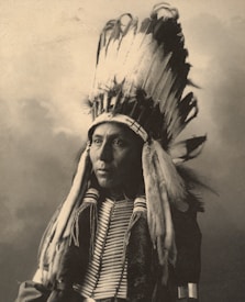 A person wearing a traditional Native American headdress with long feathers. The attire includes decorative elements and a beaded necklace. The image has a sepia tone, giving it an antique appearance.
