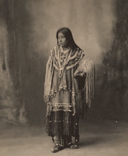 A person stands wearing traditional, intricately beaded clothing with long fringes. The garment is adorned with numerous strings of jewelry and patterns. The backdrop is a soft, blurred texture that complements the sepia tone of the photograph.