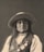 A portrait of a person wearing a wide-brimmed hat, adorned with long hair and various medals and beads. The expression is serious, evoking a sense of dignity and presence.
