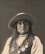 A portrait of a person wearing a wide-brimmed hat, adorned with long hair and various medals and beads. The expression is serious, evoking a sense of dignity and presence.