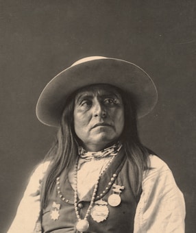 A portrait of a person wearing a wide-brimmed hat, adorned with long hair and various medals and beads. The expression is serious, evoking a sense of dignity and presence.