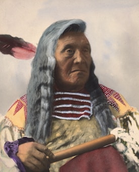 An individual with long grey hair is wearing traditional attire with intricate beadwork and holding a wooden stick adorned with feathers. The clothing features colorful patterns and a feather is also visible in the hair, suggesting cultural significance.